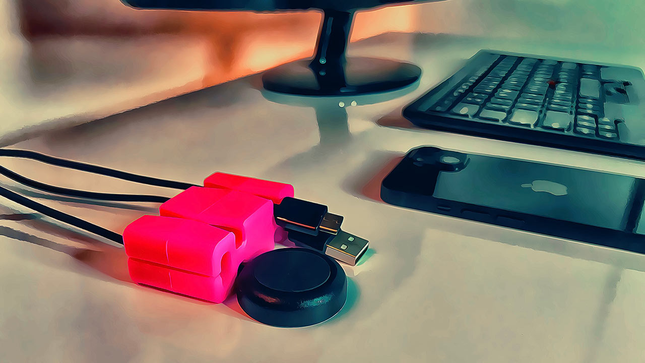 One pink CordBrick Desktop Organizer Holding three cords in the front left foreground - an Apple iPhone and keyboard and monitor on a white desk.