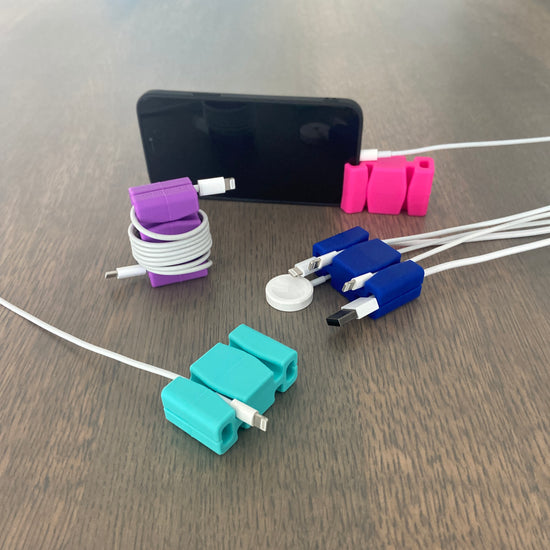 Four CordBricks, clockwise from bottom, an aqua one holding one cord, a purple one wrapping one phone charger cable, a pink one standing a phone, and a dark blue one holding several cords.
