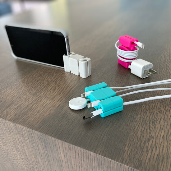 Three CordBricks shown - an aqua one in the foreground holding four cords, a light gray one on the top left standing a phone, and a pink one with one cord wrapped on the top right. 